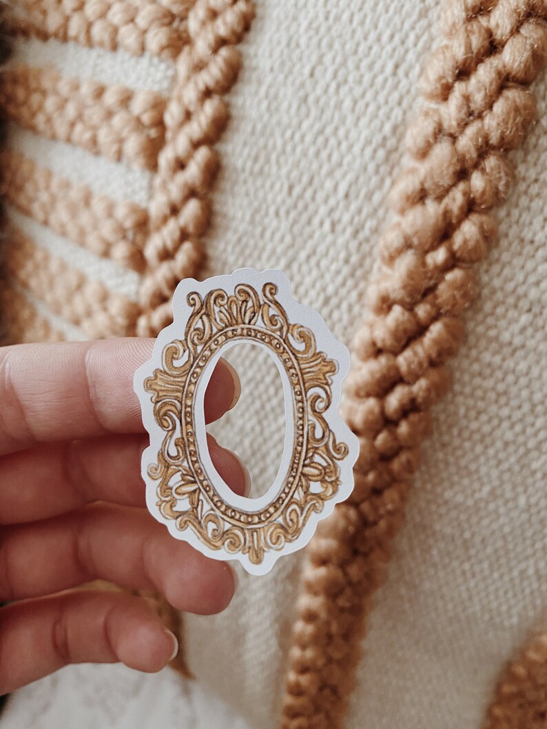 May include: A sticker featuring a gold-colored, ornate oval frame design. The frame has intricate scrollwork and detailing, with a white background. The sticker is held in a hand against a textured, beige background.
