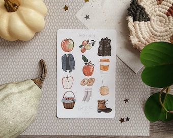May include: A sheet of stickers with an apple picking theme. The stickers feature illustrations of apples, a jacket, a cup of coffee, a basket of apples, a pair of socks, a boot, and a pie. The text on the sheet reads "Apple Picking" and "Owl & Fern by Saphire".