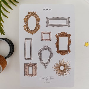 May include: A sticker sheet featuring various ornate frame designs in gold, brown, and silver. The sheet includes oval, rectangular, and sunburst frame illustrations. The text "FRAMES" and "Opal & Fern" are visible. Two rolls of decorative tape and a fern are in the background.