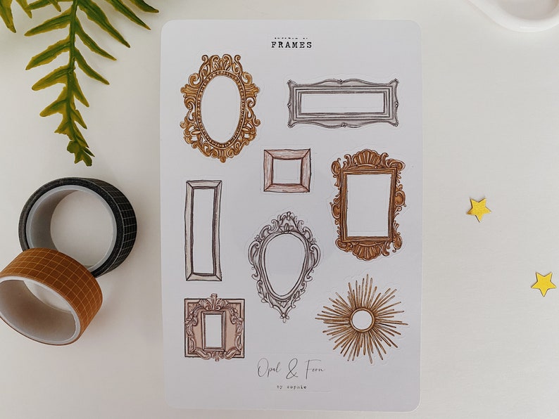 May include: A sticker sheet featuring various ornate frame designs in gold, brown, and silver. The sheet includes oval, rectangular, and sunburst frame illustrations. The text "FRAMES" and "Opal & Fern" are visible. Two rolls of decorative tape and a fern are in the background.