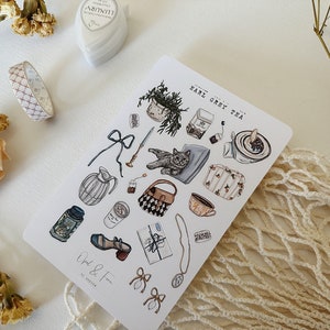 May include: A sheet of stickers featuring illustrations of a cat, teacups, a teapot, a potted plant, a purse, and other objects. The stickers are on a white background and have a light blue, brown, and white color scheme. The text "Earl Grey Tea" is written on the sheet.
