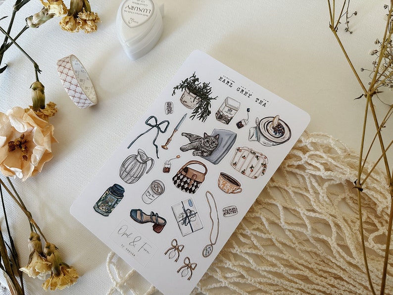 May include: A sheet of stickers featuring illustrations of a cat, teacups, a teapot, a potted plant, a purse, and other objects. The stickers are on a white background and have a light blue, brown, and white color scheme. The text "Earl Grey Tea" is written on the sheet.