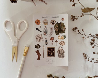 May include: A sticker sheet with autumn-themed illustrations, including pumpkins, a sweater, mushrooms, and a key. The text "AUTUMN COTTAGECORE" is at the top. A pair of scissors, a paintbrush, and a pencil are in the foreground.
