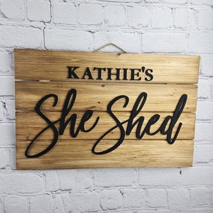 May include: Wooden sign with the text "Kathie's She Shed" in black lettering. The sign is made of light brown wooden planks and hangs from a natural twine string. The background is a white brick wall.