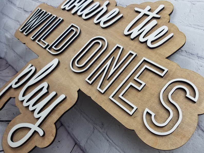 Op de afbeelding: Een houten bord met de tekst "Where the Wild Ones Play" in witte letters met zwarte contouren. Het bord is gemaakt van lichtbruin hout en heeft een rustieke uitstraling.