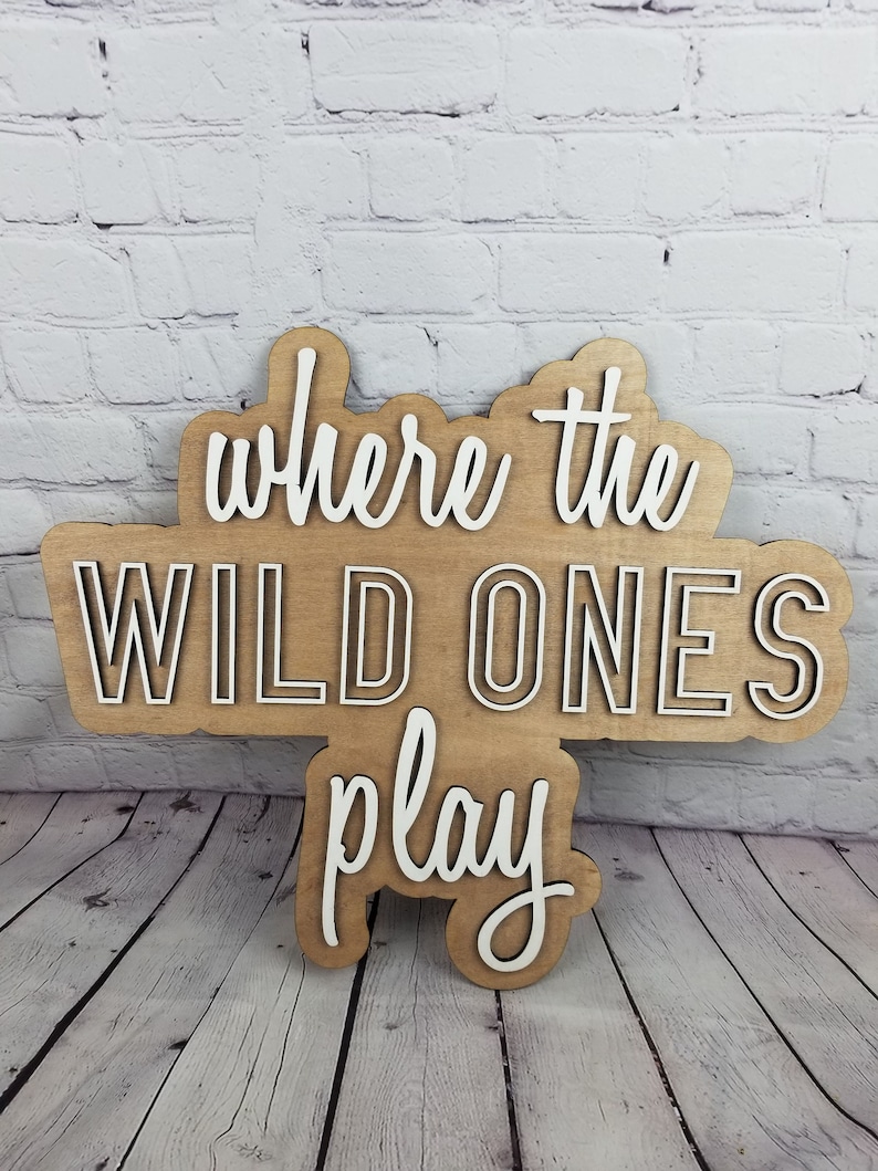 Op de afbeelding: Een houten bord met de tekst "where the wild ones play" in witte letters. Het bord is bruin en heeft een rustieke uitstraling.