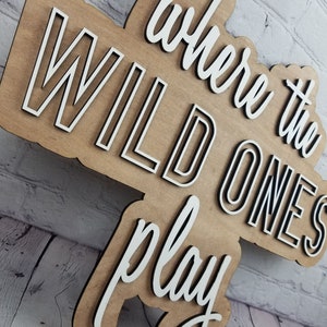 Op de afbeelding: Een houten bord met de tekst "where the wild ones play" in witte letters met zwarte contouren. Het bord is gemaakt van lichtbruin hout en heeft een rustieke uitstraling.