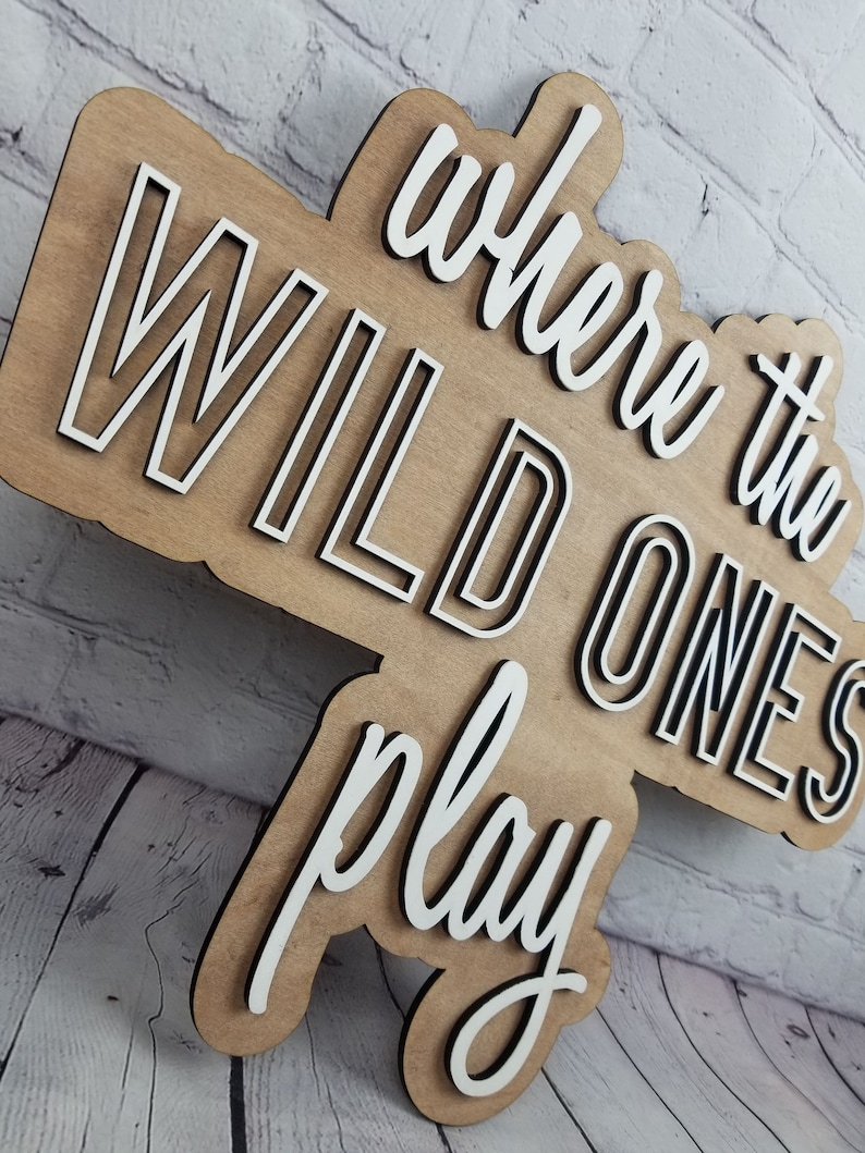 Op de afbeelding: Een houten bord met de tekst "where the wild ones play" in witte letters met zwarte contouren. Het bord is gemaakt van lichtbruin hout en heeft een rustieke uitstraling.