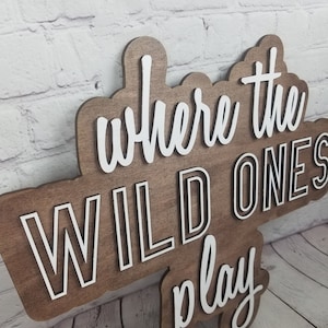 Op de afbeelding: Een houten bord met de tekst "Where the Wild Ones Play" in witte letters met een zwarte omtrek. Het bord is bruin en heeft een rustieke uitstraling.