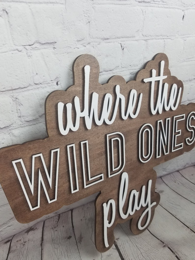 Op de afbeelding: Een houten bord met de tekst "Where the Wild Ones Play" in witte letters met een zwarte omtrek. Het bord is bruin en heeft een rustieke uitstraling.