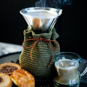 May include: A glass pour-over coffee maker with a green knitted cozy and a leather tie. Steam rises from the coffee maker. A small glass pitcher of coffee and pastries are in the foreground. The background is black.