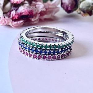 Peut inclure: Un ensemble de trois bagues empilables en argent avec une rangée de petites pierres précieuses. La bague du haut a des pierres précieuses vertes, la bague du milieu a des pierres précieuses bleues et la bague du bas a des pierres précieuses rouges.