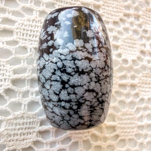 May include: A polished, barrel-shaped snowflake obsidian gemstone bead featuring a deep black base color with distinct, white, flower-like radial inclusions scattered across its smooth, glossy surface, showcasing natural volcanic glass patterns.