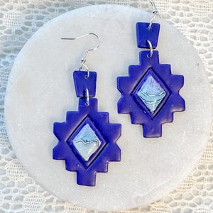 May include: A pair of handcrafted, indigo-colored earrings. Each earring features a geometric design with a central diamond-shaped inlay of white and blue. The earrings are suspended from silver-colored ear wires.
