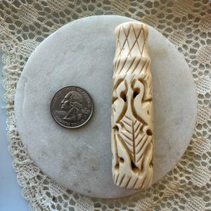 May include: A carved, off-white cylindrical object with intricate patterns, including geometric and avian designs. A U.S. quarter is placed next to the object for size comparison. The object is on a white, round stone.
