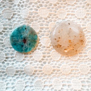 May include: Two circular pendants. The left pendant is a vibrant turquoise blue with dark veining and a small hole near the top. The right pendant is a translucent off-white with scattered brown speckles and a smooth, polished surface finish.