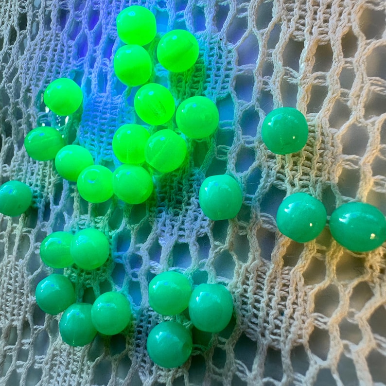以下が含まれることがあります： 明るい緑色の丸いビーズが、クリーム色のオープン織りの生地に散らばっています。ビーズは光沢のある仕上がりで、光っているように見えます。背景は青と紫の色合いのグラデーションです。
