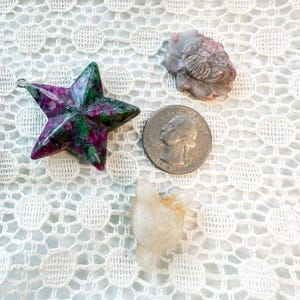May include: A selection of gemstone carvings and a quarter coin. The carvings feature a star-shaped pendant with green and magenta colours, a small bird, and a fish. The items are displayed on a white lace surface.