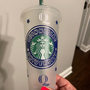May include: A clear plastic reusable cup with a blue and white design featuring the text "Quinnipiac University" and a green Starbucks logo. The cup has a white letter "Q" on the top and bottom.