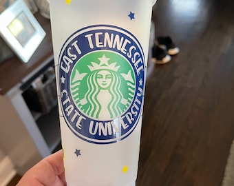 May include: A white reusable cup with a blue and green East Tennessee State University logo. The logo features a green Starbucks siren with a crown and a blue circle with the words "East Tennessee State University" around the outside. The cup is decorated with yellow stars.