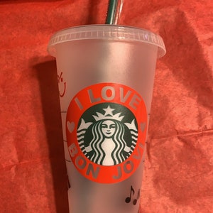 May include: A clear plastic reusable cup with a red and white design that says "I Love Bon Jovi" with a green Starbucks logo in the center. The cup has a black straw and musical notes on the bottom.