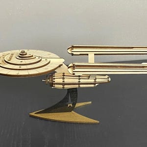 Puede incluir: Un modelo de madera de la nave espacial USS Enterprise de Star Trek. El modelo está hecho de madera cortada con láser y presenta detalles intrincados, incluida la icónica sección de platillo, las nacelas y la propulsión de deformación. El modelo está montado en un soporte de madera.
