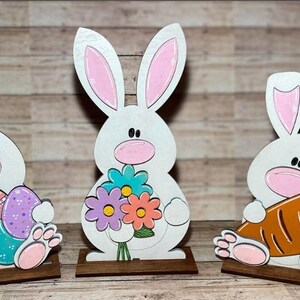 May include: Three wooden bunny figurines, each with a different Easter theme. One bunny holds colorful Easter eggs, another holds a bouquet of flowers, and the third holds a carrot. The bunnies are painted white with pink ears and noses.