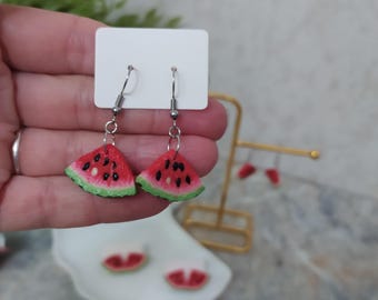 May include: Watermelon slice earrings with red, black, and green details. The earrings are on silver-colored hooks. The earrings are displayed on a white card. The earrings are a fun, summery accessory.