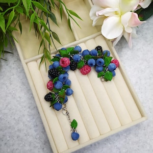 Op de afbeelding: Een bedelarmband met een tros miniatuur kunstmatige bosbessen, frambozen en bramen, afgewisseld met kleine groene blaadjes. De fruitbedels zijn bevestigd aan een zilverkleurige metalen ketting met een bungelende bessenbedel aan het uiteinde.