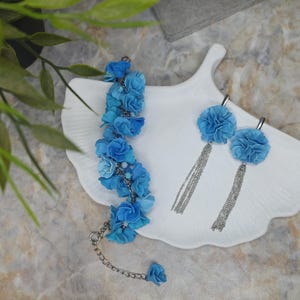 May include: A set of blue flower jewellery, comprising a bracelet and a pair of earrings. The bracelet is made of blue fabric flowers strung together on a silver chain. The earrings are also made of blue fabric flowers with silver chains hanging from them.