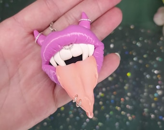 May include: A purple, glossy lips-shaped pendant with fangs and a tongue sticking out. The pendant has silver piercings on the tongue, horns, and lip. The teeth are white, and the tongue is a light peach color. The pendant is held in a hand.