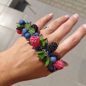 May include: A handmade ring adorned with a collection of colourful berries. The ring displays bright red raspberries, deep blue blueberries, and dark blackberries, with green leaves. The berries are attached to a silver chain.