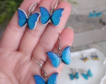 May include: A collection of butterfly earrings in various styles. The earrings feature vibrant blue butterfly wings with black detailing and white borders. The earrings are made of metal and are displayed on a hand and a white plate.