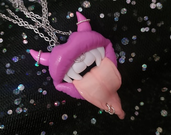 May include: A pink polymer clay necklace with a detailed mouth, fangs, and tongue. The necklace features silver hoop earrings on the mouth and tongue.