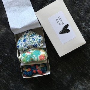 Könnte beinhalten: Eine weiße Box mit dem Text "LOVE BOX" und einem schwarzen Herzen auf dem Deckel. Die Box enthält drei kleine herzförmige Kissen mit Blumenmustern in Blau, Grün und Rot.