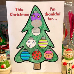 May include: A Christmas-themed drawing of a green Christmas tree with colorful ornaments. The text reads "This Christmas I'm thankful for..." with the words "My friends" on one of the ornaments. Two snowman figurines are on either side.