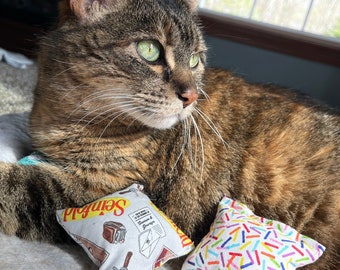 May include: A tabby cat with green eyes is lying on a white blanket with two cat toys. One toy is a square pillow with a colorful sprinkle pattern. The other toy is a square pillow with a vintage-style print of a brown suitcase, a letter, and the word "Sendall".