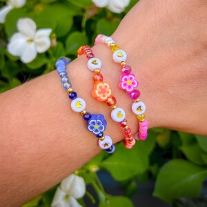 Könnte beinhalten: Drei farbige Perlenarmbänder mit Blumenanhängern. Die Armbänder sind an einem Handgelenk übereinander gelegt. Die Anhänger sind blau, orange und pink.