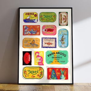 Puede incluir: Un cuadro enmarcado con un collage de etiquetas de latas de sardinas vintage. Las etiquetas rectangulares presentan varios colores, como verde, amarillo, rojo y azul, con texto e ilustraciones. La impresión se muestra sobre un suelo de madera.