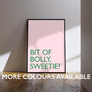 May include: A framed pink poster with the text "BIT OF BOLLY, SWEETIE?" in green. Below the text is the phrase "MORE COLOURS AVAILABLE" in white. The poster is displayed on a wooden floor.