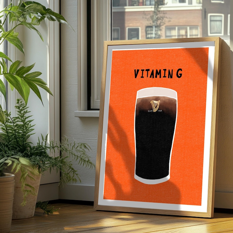 Peut inclure: Impression d'art encadr&eacute;e repr&eacute;sentant une pinte de stout fonc&eacute; avec une t&ecirc;te blanche, sur fond orange. Le texte "VITAMIN G" est affich&eacute; au-dessus de la pinte. L'&oelig;uvre est dans un cadre en bois, appuy&eacute;e contre une fen&ecirc;tre.