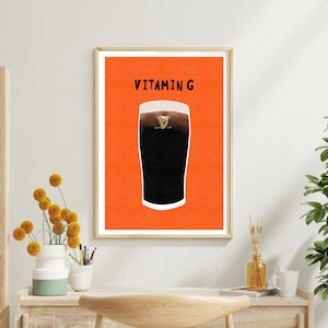 Peut inclure: Une affiche encadr&eacute;e repr&eacute;sentant une pinte de bi&egrave;re noire avec une t&ecirc;te cr&eacute;meuse, sur fond orange vif. Les mots "VITAMIN G" sont imprim&eacute;s au-dessus de la bi&egrave;re. L'&oelig;uvre est dans un cadre dor&eacute;.