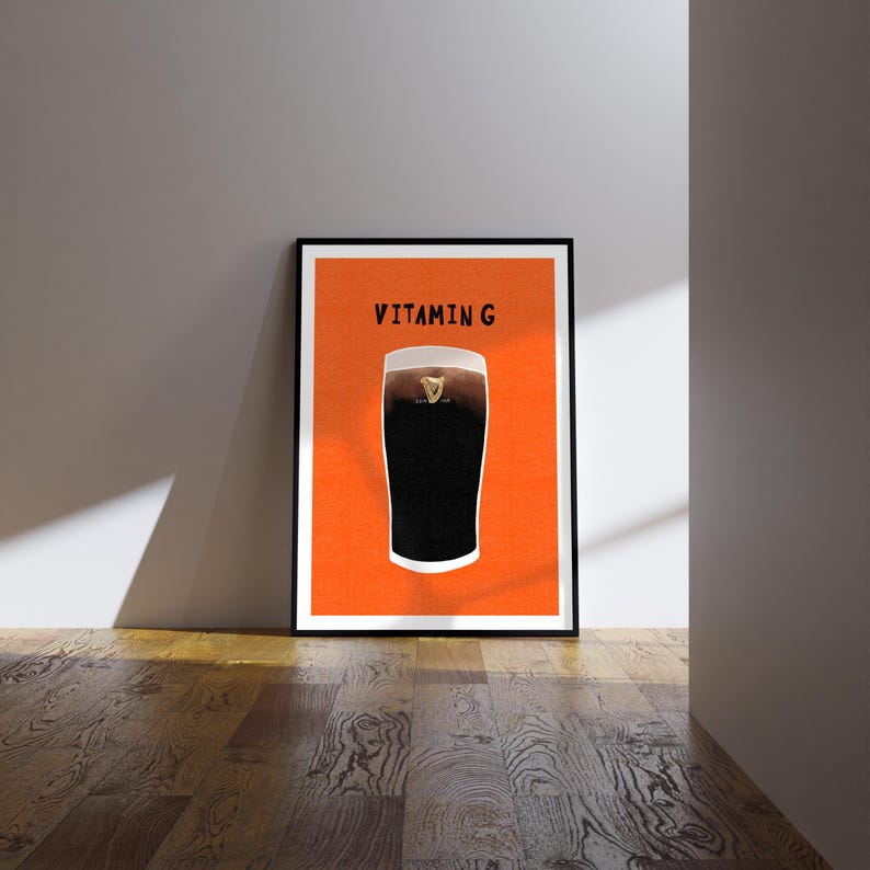 Peut inclure: Une affiche encadr&eacute;e repr&eacute;sentant une pinte de stout fonc&eacute; sur fond orange. Le texte "VITAMIN G" est imprim&eacute; au-dessus de la pinte. L'affiche est dans un cadre noir et est pos&eacute;e contre un mur blanc sur un sol en bois.