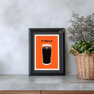 Peut inclure: Impression encadr&eacute;e repr&eacute;sentant une pinte de stout fonc&eacute; avec une t&ecirc;te cr&eacute;meuse, sur fond orange. L'impression est intitul&eacute;e "Vitamin G" en caract&egrave;res gras, dans un cadre noir. L'&oelig;uvre est expos&eacute;e sur un comptoir gris.