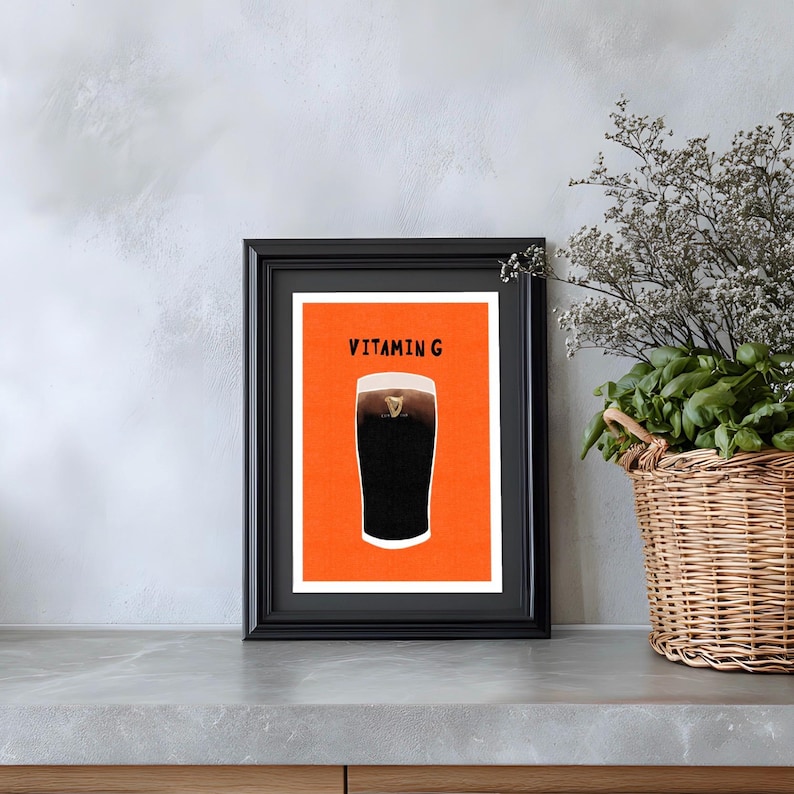 Peut inclure: Impression encadr&eacute;e repr&eacute;sentant une pinte de stout fonc&eacute; avec une t&ecirc;te cr&eacute;meuse, sur fond orange. L'impression est intitul&eacute;e "Vitamin G" en caract&egrave;res gras, dans un cadre noir. L'&oelig;uvre est expos&eacute;e sur un comptoir gris.
