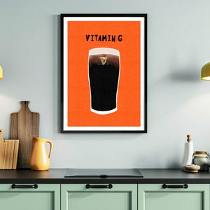 Peut inclure: Impression d'art encadr&eacute;e repr&eacute;sentant une pinte de stout fonc&eacute; sur fond orange. Le texte "VITAMIN G" est affich&eacute; au-dessus de la pinte. L'impression est dans un cadre noir et est accroch&eacute;e &agrave; un mur bleu clair.
