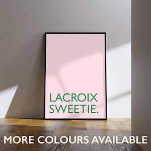 May include: A framed pink art print with the words "LACROIX SWEETIE" in green. The phrase "MORE COLOURS AVAILABLE" is printed at the bottom. The print is displayed on a wooden floor against a white wall.