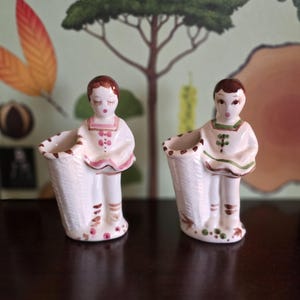May include: Two vintage ceramic figurines of children holding baskets. The figurines are painted with white, pink, and brown details. The children have closed eyes and are wearing dresses. The baskets are textured and appear to be woven.