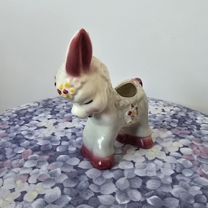 Puede incluir: Una maceta de cerámica vintage con forma de llama, en crema, con detalles burdeos en las orejas, pezuñas y detalles florales. La llama tiene una pequeña abertura en la espalda para plantar. La maceta está sobre una superficie floral.