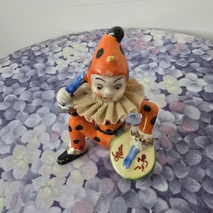 May include: A vintage ceramic clown figurine in orange with black polka dots, a ruffled collar, and a black and gold hat. The clown is holding a yellow and green musical instrument with blue and red accents.